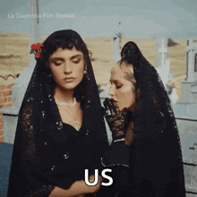two women wearing black veil and gloves are standing next to each other in a cemetery and one of them is saying us