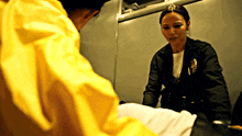 a woman in a black jacket with a badge on her chest talks to a man in a yellow jacket
