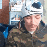 a young man wearing a beanie and a camo jacket is sitting in a room .