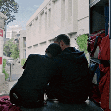 two men sit on the back of an ambulance with their heads on each other looking out