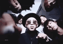 a group of young men wearing hats and sunglasses are standing around a man wearing a hat and sunglasses .