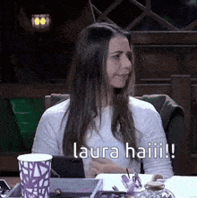 a woman is sitting at a table with a cup of coffee and a cell phone and says `` laura haiii ! ''