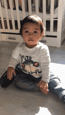 a little boy wearing a shirt that says kap sits on the floor in front of a crib