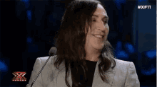 a man with long hair is smiling in front of a microphone with #xf11 written on the bottom right