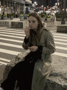 a woman in a trench coat sits on a rock eating food