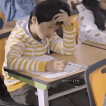a young boy is sitting at a desk in a classroom with his hands on his head .