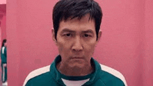 a man in a green jacket is standing in front of a pink wall and looking at the camera .