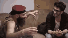 two men are sitting on a couch talking to each other . one of the men is wearing a red hat and glasses .