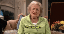 an elderly woman in a green jacket is sitting on a couch and says what the hell .