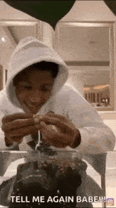a man in a white hoodie is sitting at a table with a cake and says tell me again babe !!