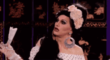 a drag queen with a flower in her hair is holding a piece of paper in her hand .