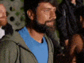 a man with a beard wearing a blue shirt and a green jacket is standing in a crowd of people .