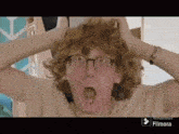 a young man with curly hair and glasses is holding his head with his hands and making a surprised face .
