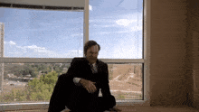 a man in a suit and tie sits on the floor in front of a large window