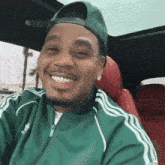 a man wearing a green adidas jacket and a green hat is smiling while sitting in a car .