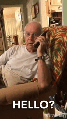 an elderly man is sitting on a couch talking on a cell phone and saying hello .