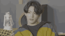 a young man in a yellow and blue jacket is sitting in a room with a lantern in the background .