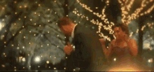 a man and a woman are dancing in a park at night with christmas lights in the background .