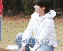a man in a white hoodie sits on the grass with korean writing on the ground