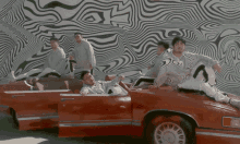 a group of men are sitting in a red car in front of a wall with a striped pattern