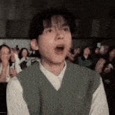 a young man wearing a green vest and a white shirt is making a funny face in front of a crowd .