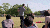a group of people are sitting on the grass in a field watching a man standing in the middle of the field .