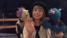 a woman with green hair is holding a stuffed animal and waving at a sesame street character .