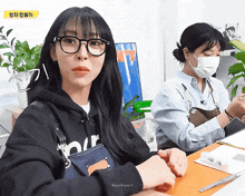 a woman wearing glasses sits at a table next to another woman wearing a face mask