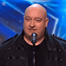 a bald man in a black jacket is singing into a microphone with a star in the background