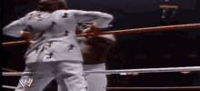 a man in a white suit is standing in a wrestling ring with another man .