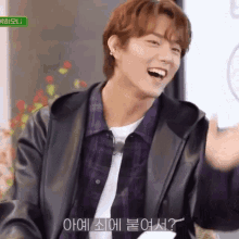 a young man wearing a leather jacket and a plaid shirt is laughing and waving his hand .
