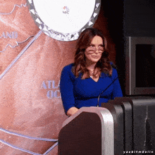 a woman in a blue dress stands at a podium in front of a wall that says nbc