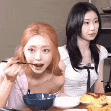 two women are sitting at a table eating food with chopsticks .