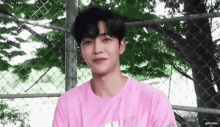 a young man wearing a pink t-shirt is standing next to a chain link fence .