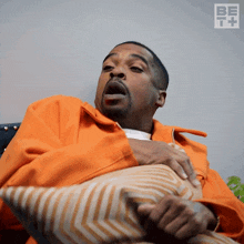 a man in an orange jacket is sitting on a couch holding a striped pillow
