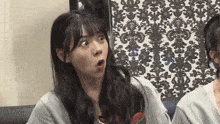 a woman is making a surprised face while sitting next to another woman in front of a wall with a pattern .