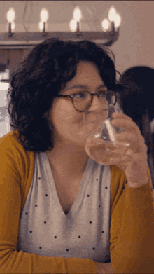 a woman wearing glasses and a yellow sweater is drinking from a glass