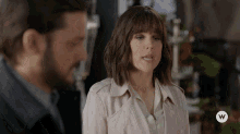 a woman in a trench coat stands next to a man in a suit with a w on the bottom right corner