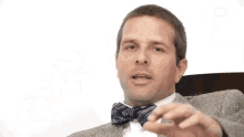 a man in a suit and bow tie is sitting in a chair and making a gesture .