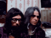 a man with long hair and glasses stands next to another man with long hair