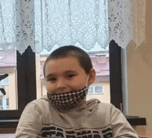 a young boy wearing a face mask is sitting at a desk in front of a window .