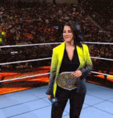 a woman in a yellow jacket and black pants is standing in a wrestling ring holding a microphone .