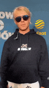 a man wearing sunglasses and a hoodie is standing in front of a blue background .
