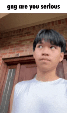 a young man in a white tank top is standing in front of a brick building and says gng are you serious .