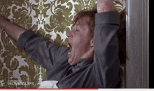 a man is yawning while standing in front of a wall with a floral wallpaper .