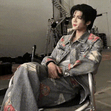 a young man in a denim jacket is sitting in a chair with his arms crossed .
