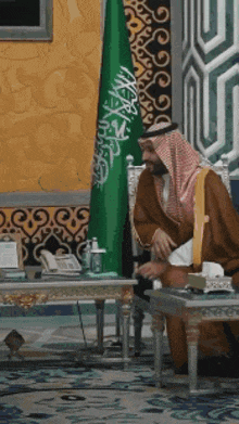 a man sitting in front of a green and white flag with arabic writing