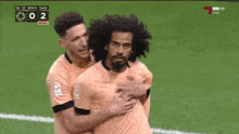 two soccer players are celebrating a goal with the scoreboard showing 0 to 2