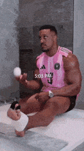 a man in a pink adidas shirt is sitting on a bathtub holding a ball