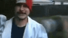 a man wearing a red hat and a white coat is standing in front of a car .